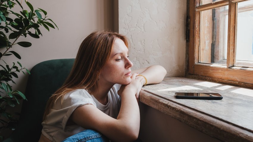 Upset redhead teen girl sitting by window looking at phone waiting call from boyfriend, feeling sad and depressed teenager looking at smartphone wait for message. Social Media depression in teens