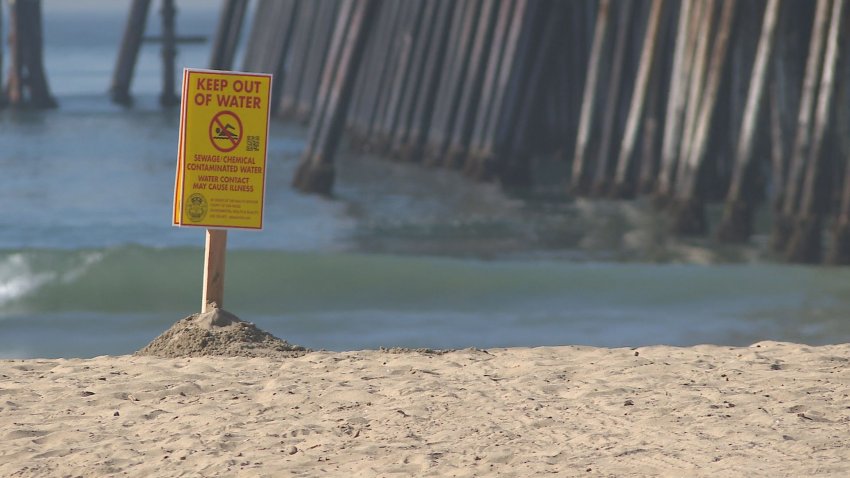 Yellow sign on a beach with waves and a pier in the background.